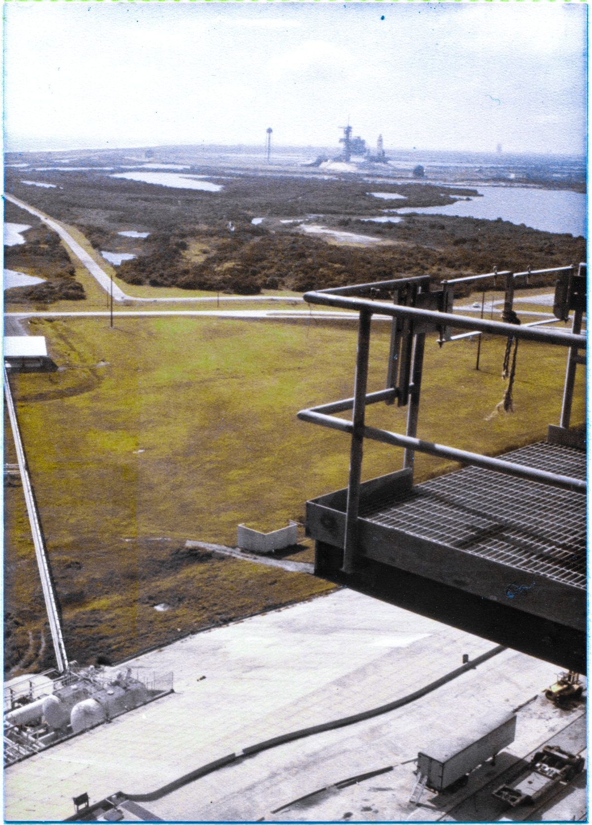 Image 039. Standing on the steel-bar grating at the 220’ elevation level, looking south southeast from a viewpoint immediately west of the Main Framing Column at the southeast corner of the Fixed Service Structure at Launch Complex 39-B, Kennedy Space Center, Florida. In the right side of the frame, part of a crossover catwalk, also at elevation 220’, can be seen. This catwalk provides access to the roof of the Rotating Service Structure when it is in the mated position. Visible on the crossover catwalk, a Safety Gate guards the sheer drop to the Pad Deck which exists when the RSS is in the demate position as it is in this image. In the distance, the Space Shuttle Columbia can be seen, carried by the Mobile Launch Platform, near the top of the Pad Slope at Pad 39-A, nearing the silhouette of the demated RSS on A Pad, completing its very slow rollout journey which started inside the VAB in predawn darkness. This is only the second time a Space Shuttle has been rolled to a launch pad, and when it flies, it will be the very first time ever that a Space Shuttle gets reused, thus validating the initial premise of reusable spacecraft which the Shuttle embodies.  Photograph by James MacLaren.